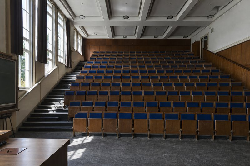 Anfiteatro universitário vazio, com filas de cadeiras de madeira e estofo azul a subir em degraus e janelas altas de um dos lados.