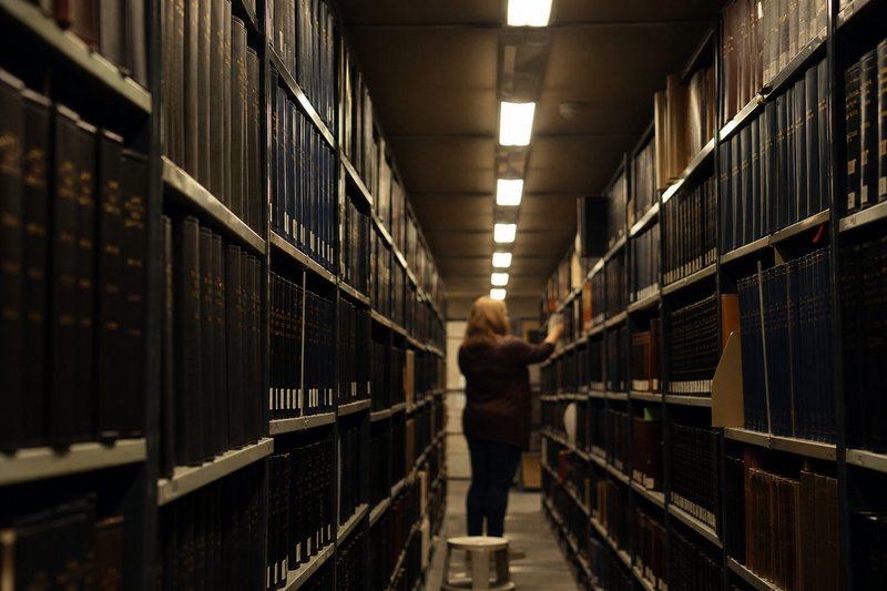 Corredor estreito entre estantes altas de biblioteca, cheias de volumes encadernados de revistas académicas.