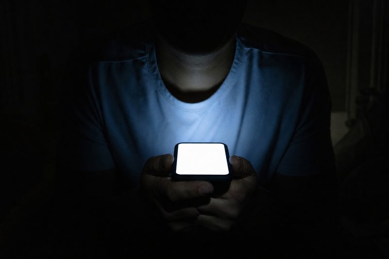 Quarto às escuras iluminado apenas pela luz azulada de um telemóvel levantado, com a figura em contraluz.