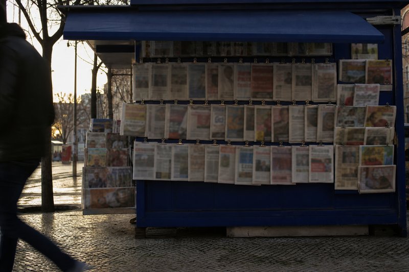 Quiosque de jornais numa rua de Lisboa ao início da manhã, com a estrutura pintada de azul e as capas expostas em fila.